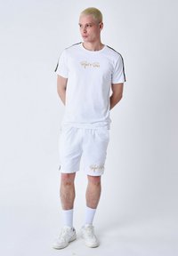 Conjunto de camiseta de algodón blanca y pantalones cortos con logotipo dorado, detalles laterales negros, cuello redondo y corte relajado. Lleva consigo unas zapatillas blancas y calcetines.
