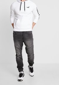 Vit hoodie med svarta dragsnören och logo, i kombination med svarta avsmalnande jeans med texturerade paneler, och svarta och vita sneakers.