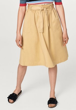 Kvinde iført en beige nederdel med højtaljet talje, bindebånd foran og påsyede lommer, sorte sandaler med flæser og en stribet top med røde kanter.