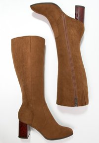 Bottes hauteur genou en daim marron avec une fermeture éclair sur le côté et un talon carré bordeaux à motif. Texture lisse avec des détails de couture minimaux.