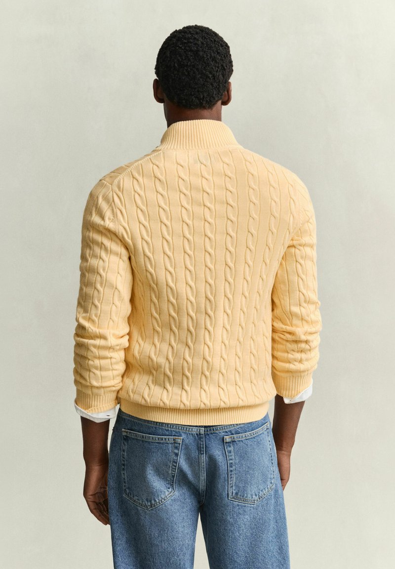 Man with short curly hair wears yellow cable-knit sweater and blue jeans, standing facing a plain light-colored background.
