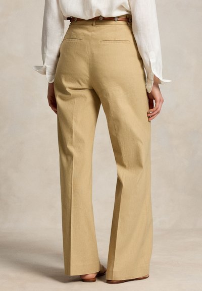 Άτομο που φοράει μπεζ φαρδιά παντελόνια με ψηλή μέση, πίσω τσέπες και καφέ ζώνη, σε συνδυασμό με ένα λευκό μακρυμάνικο πουκάμισο.