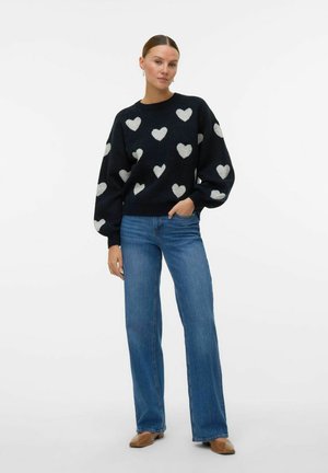Marinblå tröja med ljusgrå hjärtmönster, rundad halsringning och ballongsärmar; tillsammans med blå utsvängda jeans och bruna loafers.