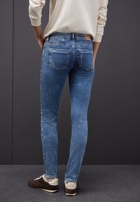 Jeans de denim azul ajustados con un acabado desgastado, cinco bolsillos y una cintura de altura media. Llevados con zapatos oscuros, mostrados desde atrás.