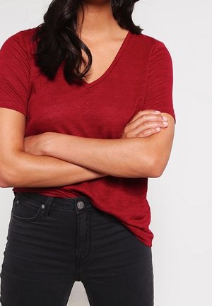 Persona con cabello largo y oscuro, vistiendo una camiseta roja de cuello en V y pantalones negros, de pie con los brazos cruzados frente a un fondo liso.