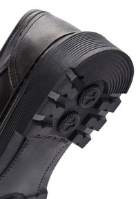 Semelle de chaussure noire avec une surface en caoutchouc texturé. Présente des crampons arrondis pour une adhérence, avec une section lisse contrastante près de la base de la chaussure.