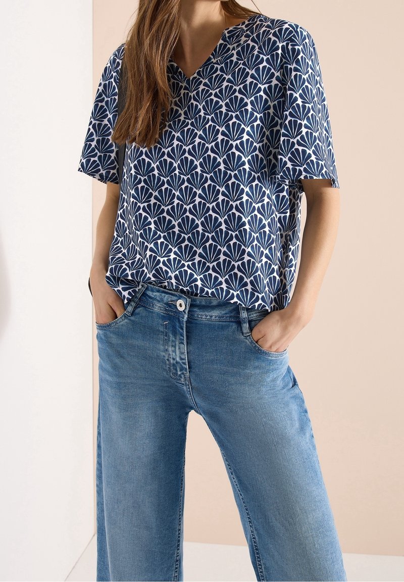 Femme portant un haut à manches courtes bleu et blanc à motifs avec un col en V, rentré dans un jean taille haute bleu clair, les mains dans les poches.