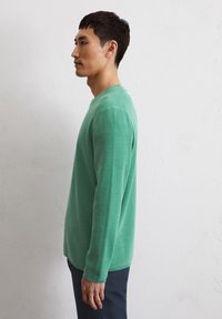 Grüner gestrickter Pullover mit langen Ärmeln und strukturiertem Finish, Rundhals-Ausschnitt und gerippten Bündchen, kombiniert mit dunklen Hosen, seitlich betrachtet.