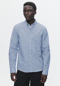 Light blue button-up shirt made of a soft fabric, featuring a chest pocket and a small embroidered logo. Long sleeves and classic collar design.