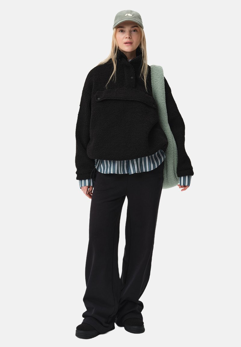 Pullover en polaire noir avec un zip sur le devant, chemise rayée en dessous, pantalons noirs et casquette verte, offrant une coupe décontractée et une texture douce.