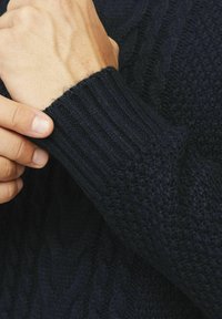 Pull en maille bleu marine avec des poignets côtelés et un motif en relief en câble. Gros plan sur une main saisissant la manche, mettant en valeur les détails du matériau.