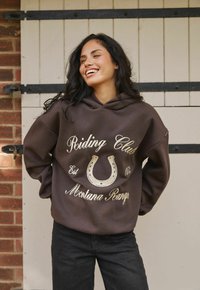 Lachende vrouw met lang donker haar, gekleed in een bruine hoodie met "Riding Club Montana Range" en een hoefijzer grafiek, staande tegen een bakstenen muur.