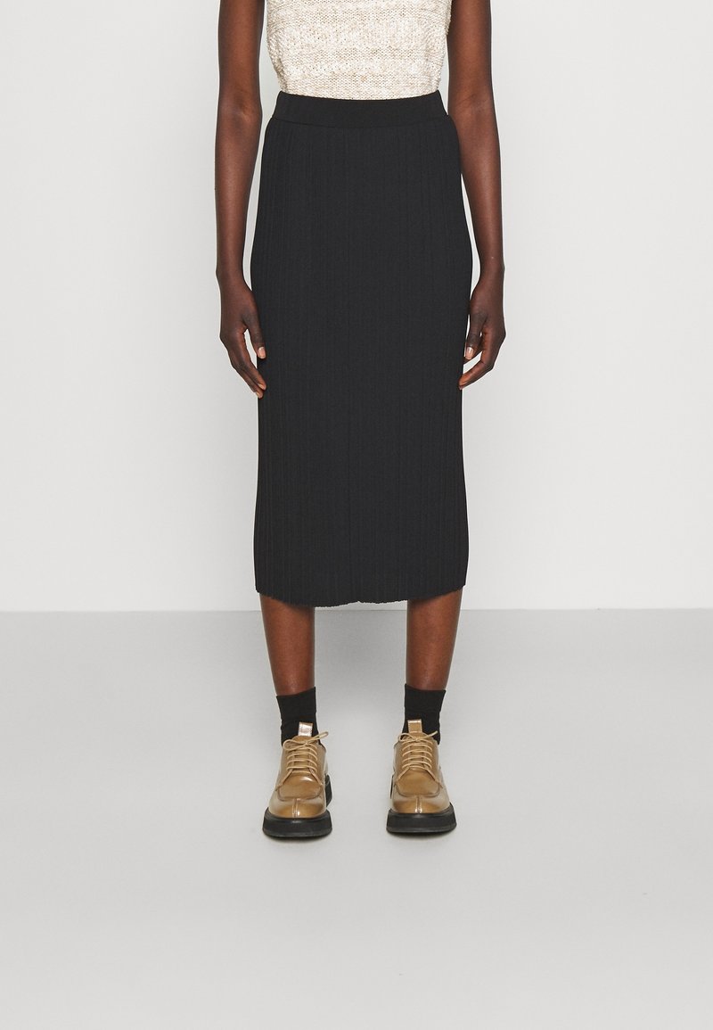 Femme portant une jupe midi côtelée noire, un haut beige sans manches, des chaussettes noires et des chaussures beige à lacets, debout devant un fond uni.
