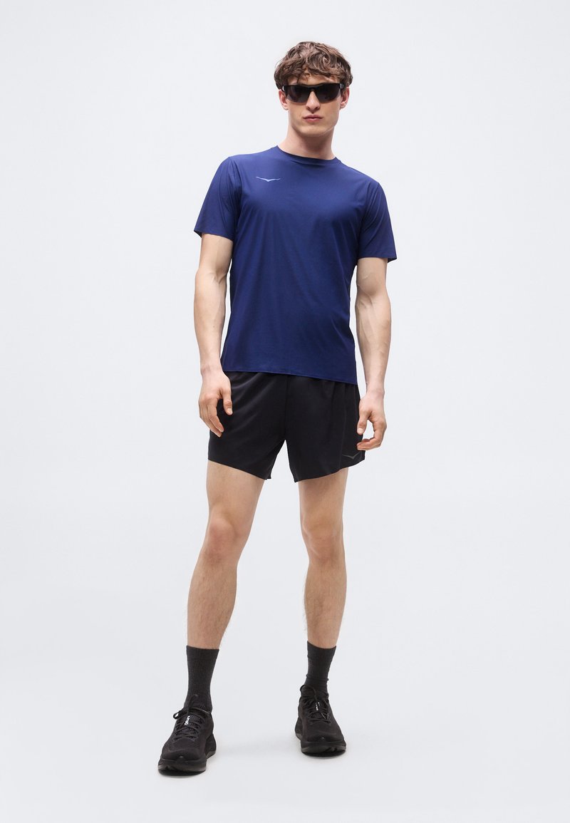 Uomo che indossa una maglietta sportiva a maniche corte blu navy, pantaloncini neri, calzini neri, scarpe da corsa nere e occhiali da sole scuri, in piedi davanti a uno sfondo semplice.