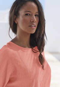 Koralfarbenes Oberteil mit lockerer Passform und weitem Ausschnitt aus weichem Material. Langes, dunkles Haar fällt locker zur einen Seite. Natürliches Make-up.