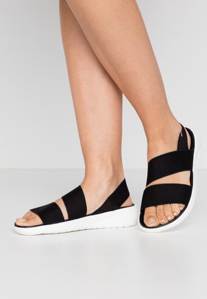 Osoba nosząca czarne sandały na elastycznych paskach z białymi podeszwami, stojąca na białej powierzchni na tle jednolitego szarego tła.