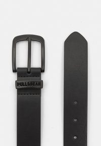 Zwarte leren riem met een rechthoekige matte zwarte metalen gesp en een logoplaat. De band heeft meerdere gaten voor verstelbaarheid.