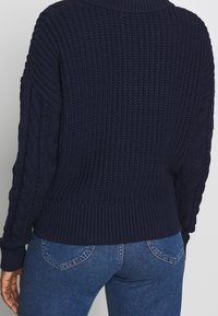 Laivastonsininen neulottu villapaita, jossa on resorikuvio, hihoissa kaapelineulekoristeet ja korkea kaulus, teksturoitu kangas ja istuva helma.