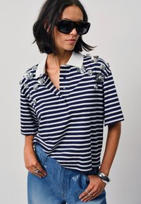 Gestreept marineblauw en wit poloshirt met decoratieve zilveren bloemenaccenten op de schouders, met een knoopsluiting aan de kraag en korte mouwen.