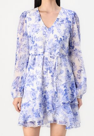 Nainen valkoisessa sifonkimekossa, jossa sininen kukkakuvio, pitkät läpikuultavat hihat, V-aukko, vyötärönauha ja kerroksittain laskostettu hame.