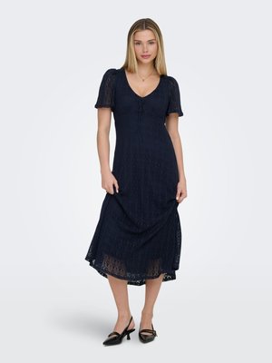 Kobieta o prostych blond włosach, ubrana w granatową koronkową sukienkę midi z bufkami i czarne buty typu slingback na niskim obcasie na jednolitym tle.