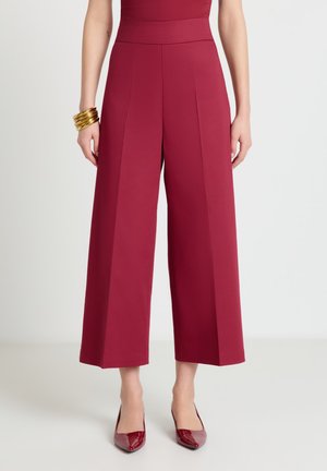 Donna che indossa pantaloni bordeaux a vita alta e gamba larga con scarpe abbinate e braccialetti d'oro al polso sinistro, in piedi su un pavimento bianco.