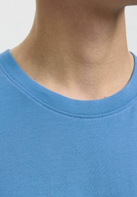 Gaiši zils kokvilnas T-krekls ar gludu tekstūru, apaļu kakla izgriezumu un smalkiem šuvju elementiem ap kaklu.