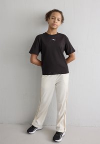 Camiseta de algodón negra con un logo blanco, combinada con pantalones color crema que tienen cremalleras laterales. Unos zapatos deportivos negros completan el conjunto.