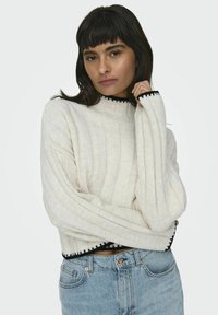 Cream ribbed sweater with a high neck and black trim. Soft texture and oversized sleeves, paired with high-waisted light blue jeans.