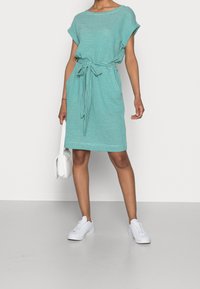 Raidallinen turkoosi mekko lyhyillä hihoilla, solmittu vyötärö ja rento istuvuus, yhdistetty valkoisiin tennareihin ja pieneen valkoiseen laukkuun.