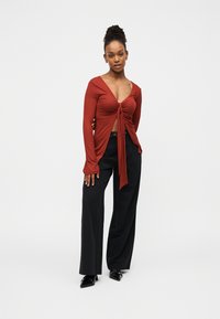 Μακρυμάνικο τοπ σε χρώμα σκουριάς με βαθύ V-λαιμόκοψη και λεπτομέρεια κόμπου μπροστά, σε συνδυασμό με φαρδιά μαύρη παντελόνα και μυτερά μαύρα τακούνια.