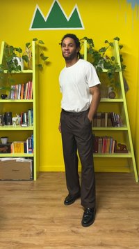 Valge T-särk, pruunid elastse vöökohaga püksid ja mustad kingad. Toad on kollaste seintega, roheliste riiulite, raamatute ja toataimedega.