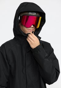 Svart vattentät jacka med huva, försedd med elastiska ärmslut. Modellen bär stora spegelglasögon med rosa och gula toner som visar kontrast.