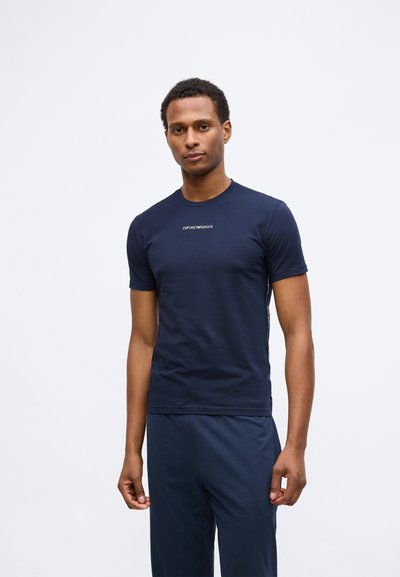 Jauns vīrietis stāv, ģērbies tumši zilos Emporio Armani T-kreklos un saskaņotās biksēs uz vienkārša baltā fona.