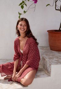 Tmavomodrý priesvitný kimonový plášť s bielymi vzormi, s previazaním v páse, krátkymi rukávmi, vrstvený cez biely top. V pozadí je kvetináč s rastlinou.