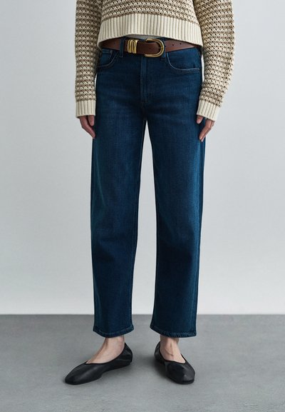 Donkerblauwe rechte jeans met een gemiddelde taillehoogte, gecombineerd met een lichtbruine riem en een beige trui met structuur. Zwarte balletschoenen maken de look compleet.
