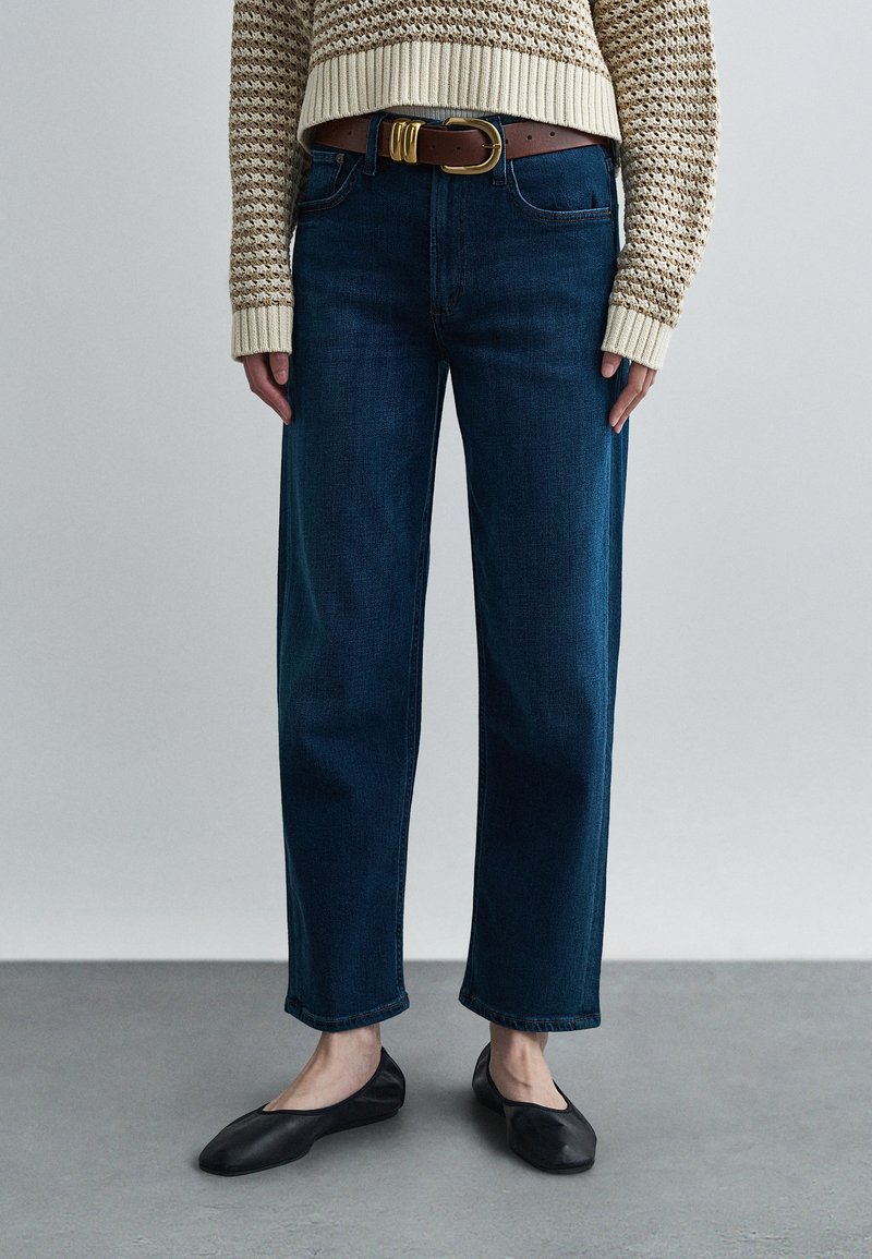 Calças de ganga azul-escuro de perna reta com cintura média, combinadas com um cinto castanho claro e um suéter texturizado bege. Sapatilhas de balé pretas completam o visual.