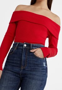 Röd ribbad off-the-shoulder topp med lång ärm, kombinerad med mörkblå högmidjade jeans som har en framficka och subtila slitningar.