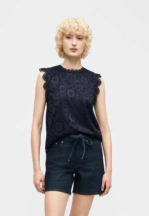 Frau mit kurzen, lockigen blonden Haaren, die ein ärmelloses marineblaues Spitzentop und dunkelblaue Jeansshorts mit gebundenem Taillenbund trägt.