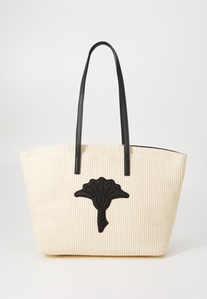 Sac fourre-tout beige en tissu tissé avec de longues poignées en cuir noir et un emblème de fleur stylisée noire centré à l'avant.