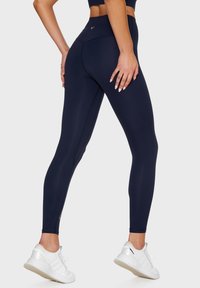 Marinblå leggings gjorda av stretchigt tyg med hög midja, med platta sömmar och diskret varumärkeslogga på ryggen. Bärs med vita träningsskor.