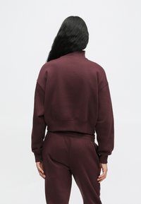 Persona con lunghi capelli neri che indossa una felpa e pantaloni della tuta color bordeaux abbinati, in piedi con le spalle rivolte verso di noi su uno sfondo semplice.