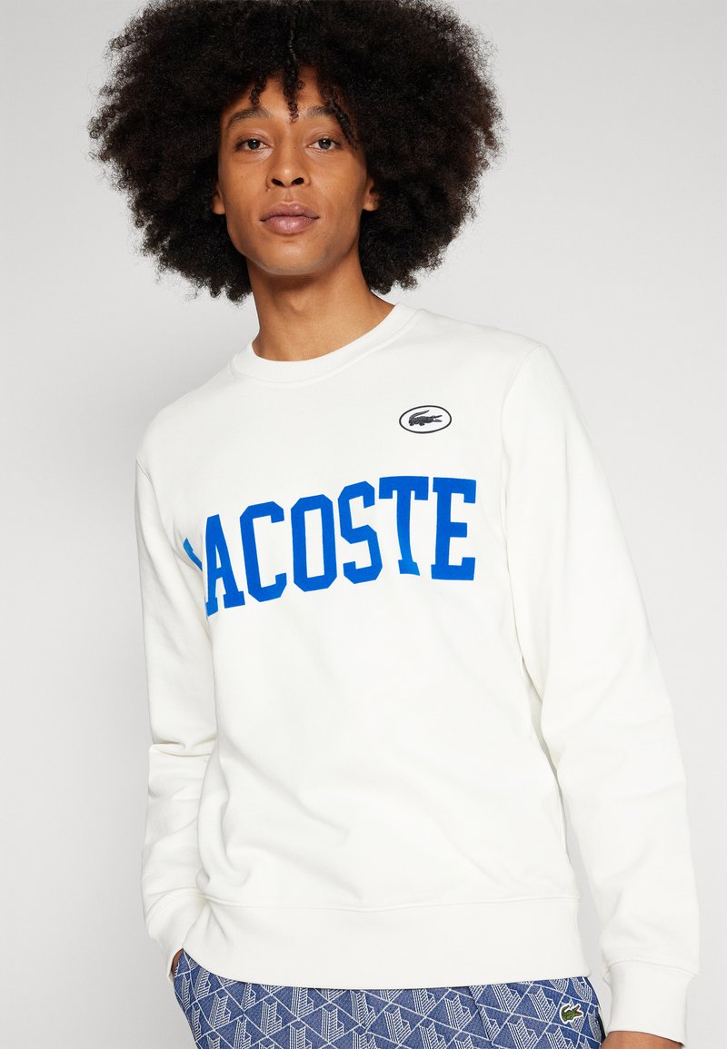 Felpa bianca con lettering blu in grassetto "LACOSTE", polsini a costine e scollatura rotonda. Include una patch nera con il logo del coccodrillo.