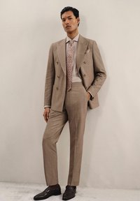 Costume beige clair à double boutonnage en tissu texturé, avec un pantalon assorti et une cravate à motifs. Des mocassins marron foncé complètent le look.