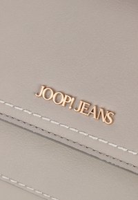 Sac en cuir gris clair avec des coutures blanches. Présente un logo en relief doré "JOOP! JEANS", ajoutant une touche subtile au design.