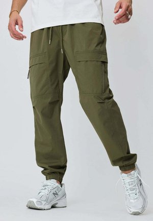 Homme portant un pantalon jogger cargo vert olive avec des poches latérales et des baskets blanches, debout les bras légèrement étendus.
