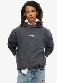 Sudadera de color gris carbón con un gran bolsillo frontal, puños acanalados y el logo "SUPERDRY" en blanco en el pecho, combinada con jeans desgastados de color azul claro.