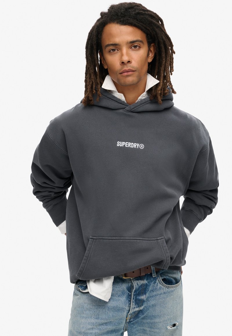 Sudadera de color gris carbón con un gran bolsillo frontal, puños acanalados y el logo "SUPERDRY" en blanco en el pecho, combinada con jeans desgastados de color azul claro.