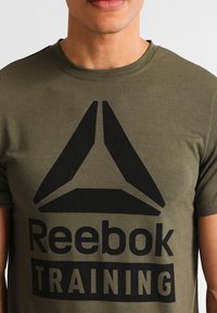 Olive green cotton t-shirt featuring a bold black graphic logo "Reebok TRAINING" at the center, round neck, and short sleeves.