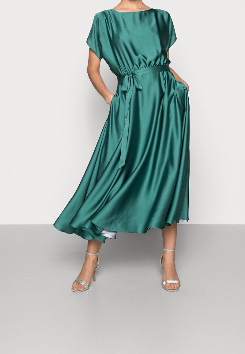 Robe en satin teal avec manches courtes, taille cintrée, poches et jupe fluide. Associée à des escarpins argentés à lanières. Texture lisse et éclat doux.